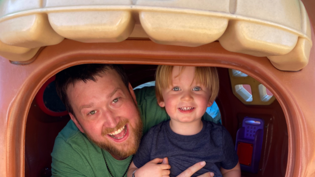 Adult and child smiling while sitting inside a playhouse at a playground.