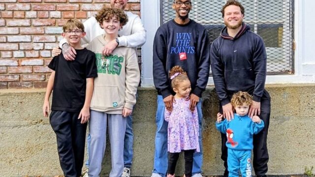 Group of six people, including two kids, posing outdoors in front of a brick wall and a window.
