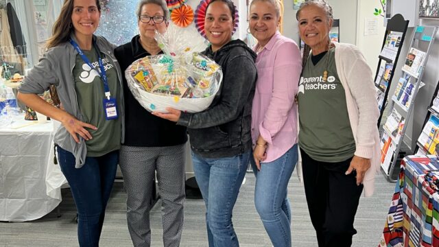 Group of people standing together indoors, one person holding a gift basket, colorful decorations in the background.
