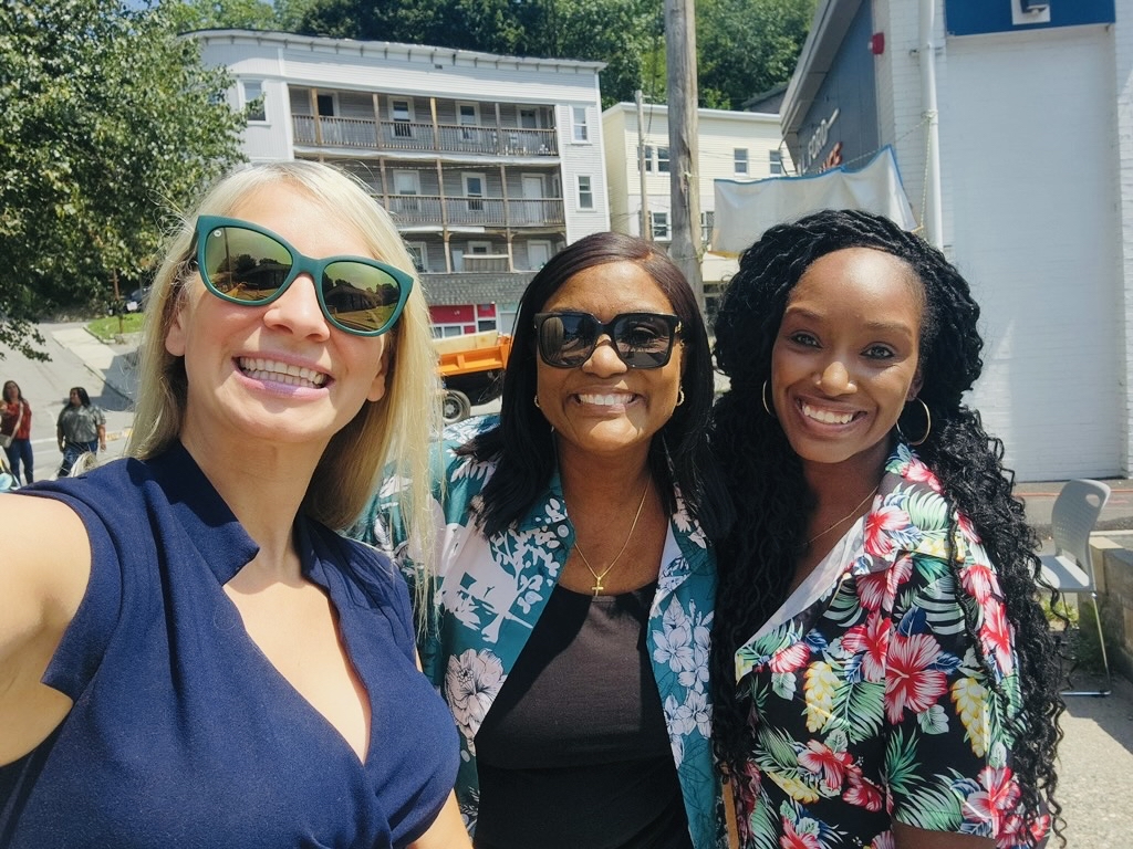 Three women smiling and posing for a selfie outdoors on a sunny day, one wearing sunglasses.