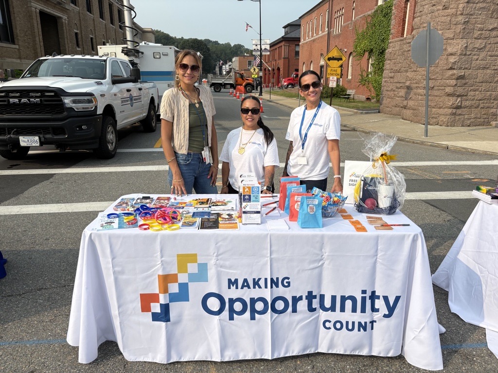 Three people standing behind a table with informational materials and branded items at an outdoor event.