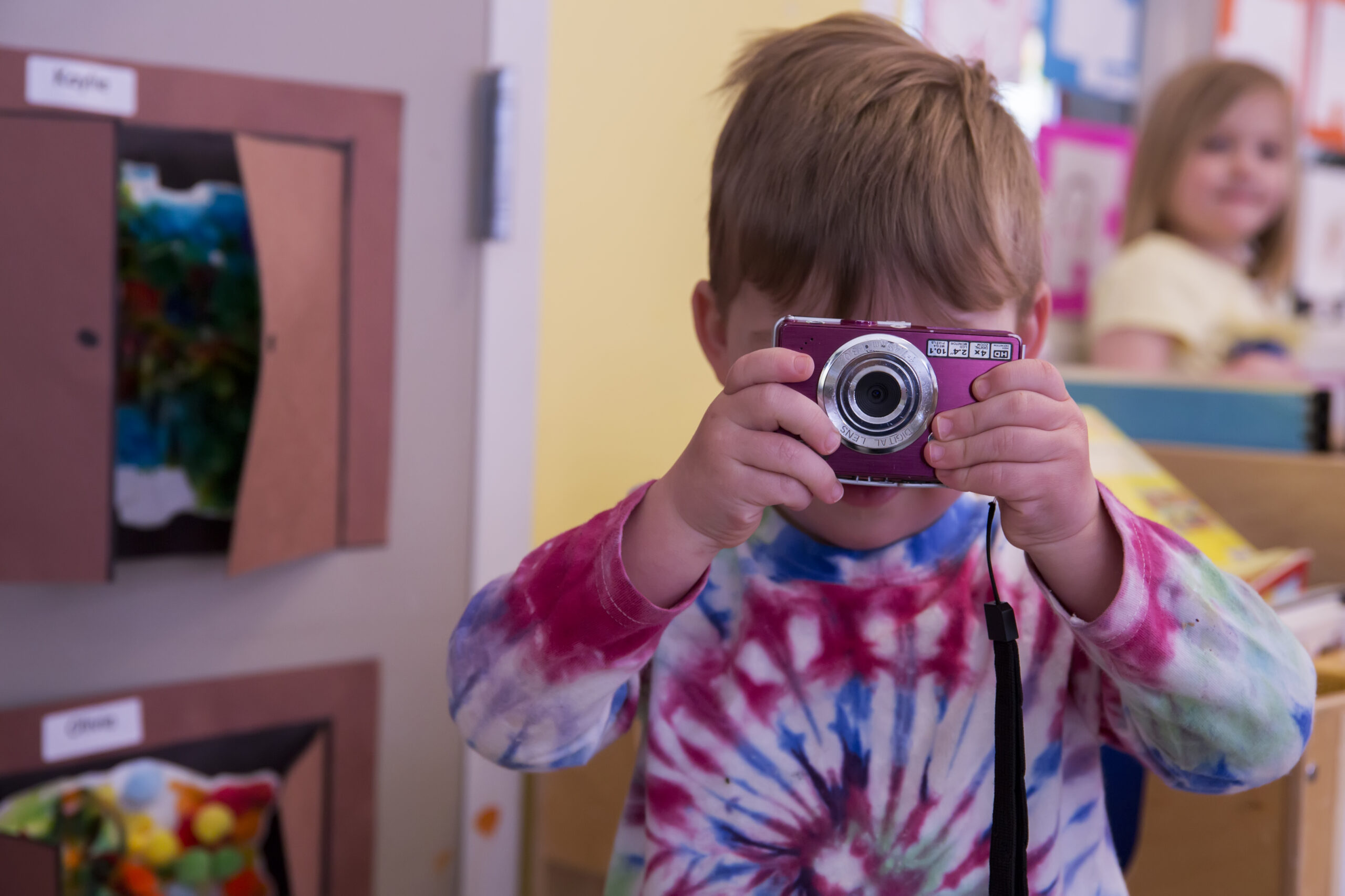 Child in a tie-dye shirt holding a camera, with a classroom setting in the background.
