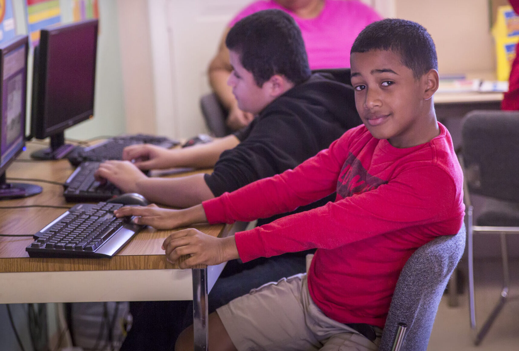 Students in a colorful classroom working on computers with a teacher in the background.