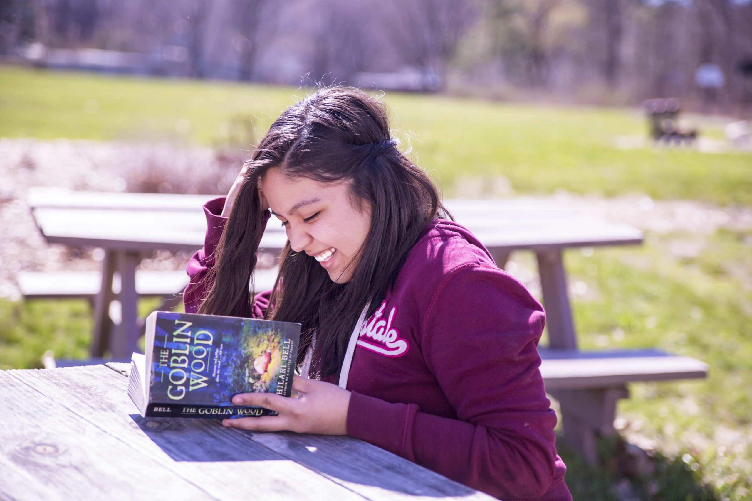 Smiling woman sitting at a picnic table, reading "The Goblin Wood" outdoors.