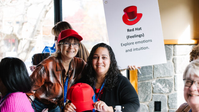 Two people holding a red hat and sign about exploring emotions at an indoor event with onlookers in the background.
