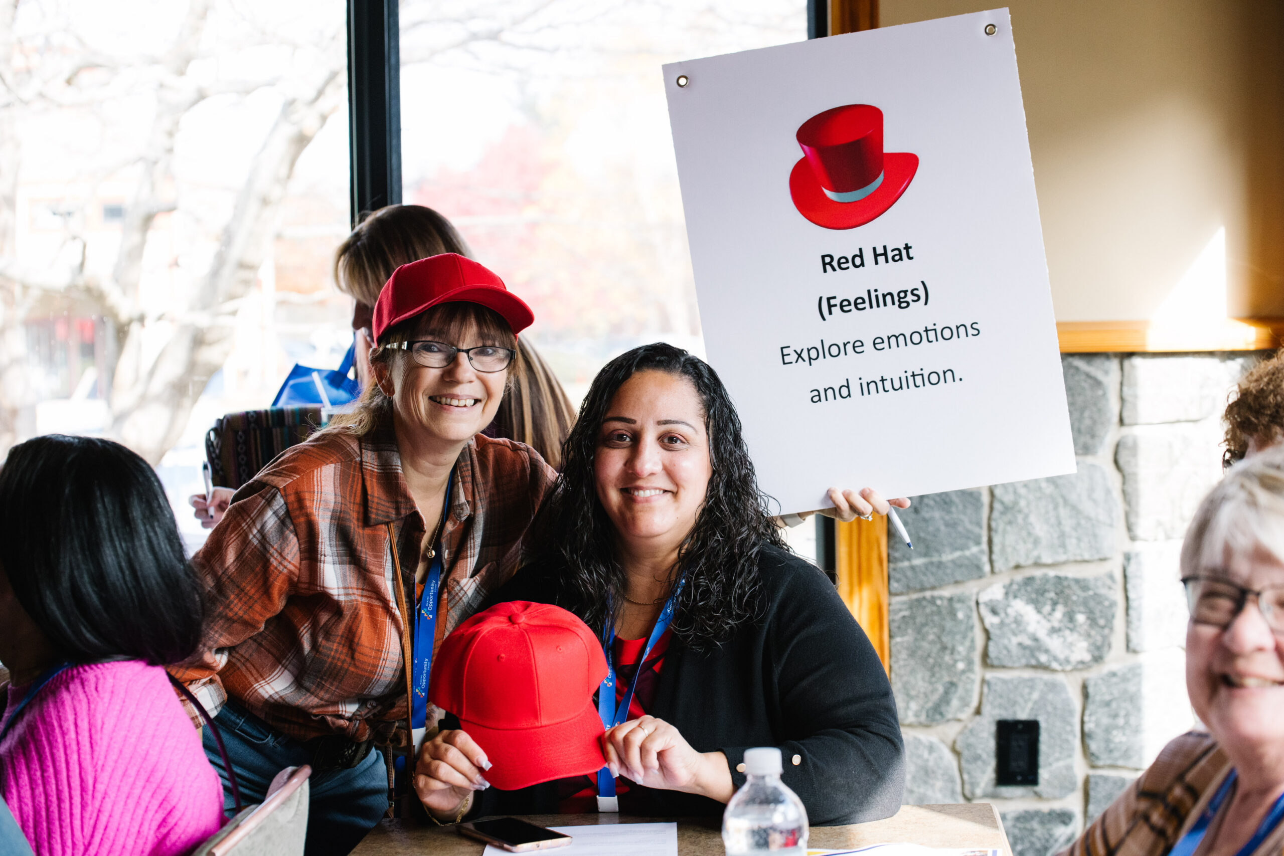 Two people holding a red hat and sign about exploring emotions at an indoor event with onlookers in the background.