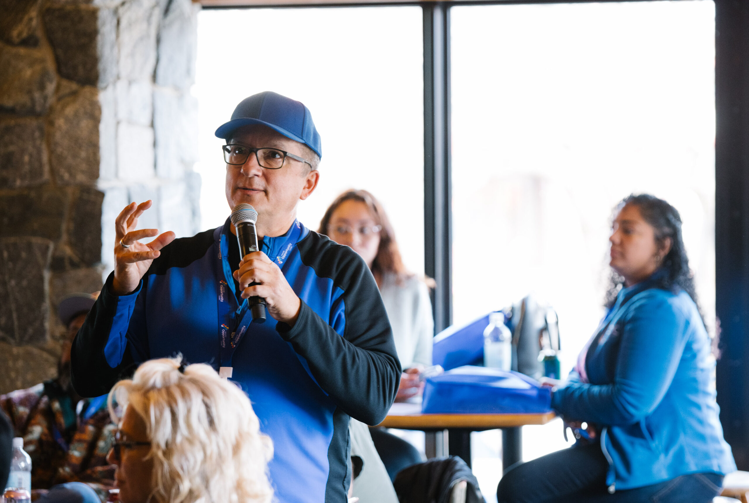 Person in blue jacket and cap speaking into a microphone in a room with seated individuals listening attentively.