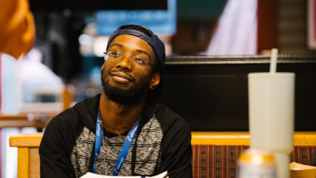 A person sitting at a table with a notebook and cup, wearing a cap and glasses, looking up with a thoughtful expression.