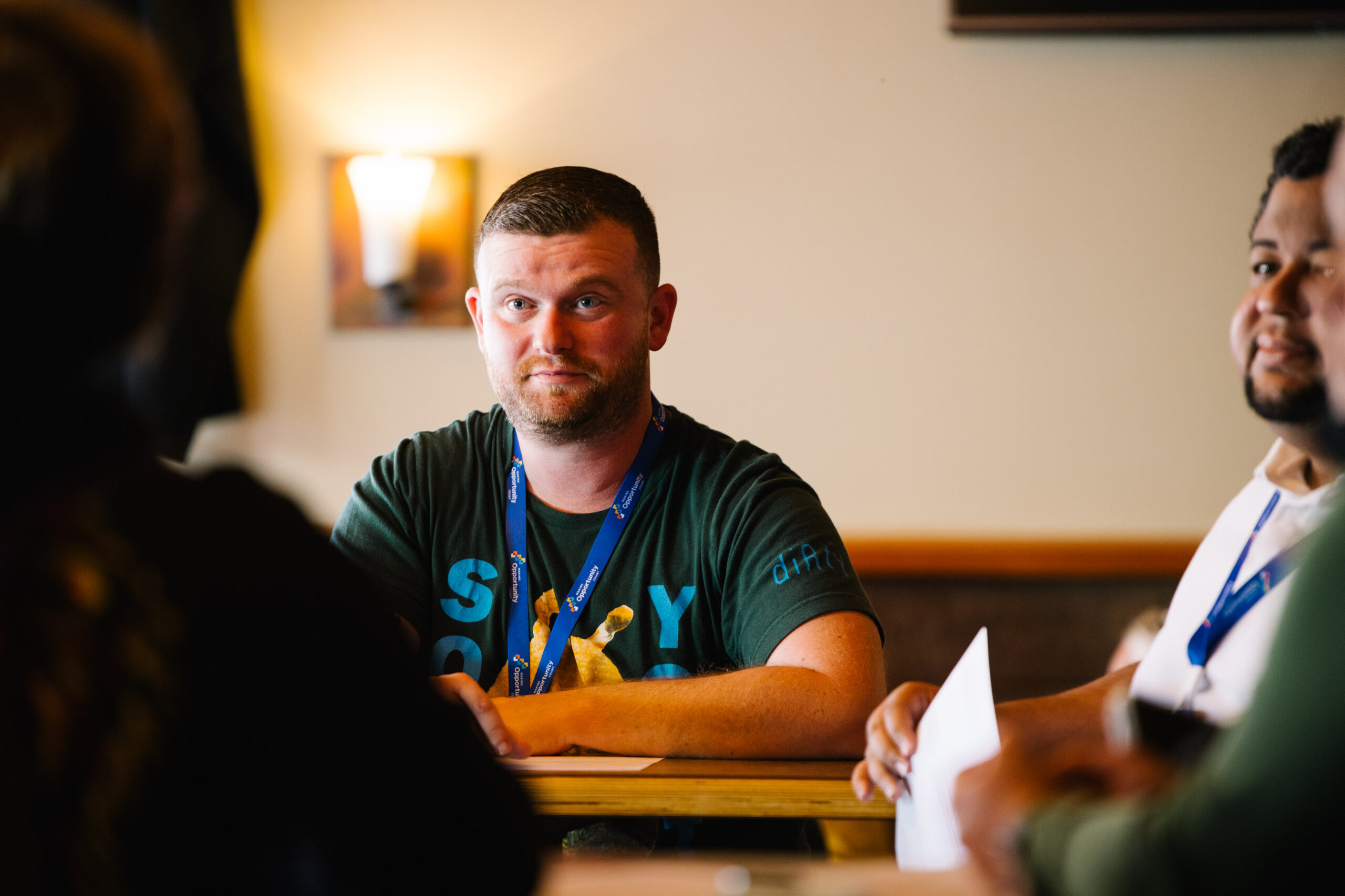 Man wearing a blue lanyard and green shirt, sitting at a table during a meeting, soft lighting in the background.