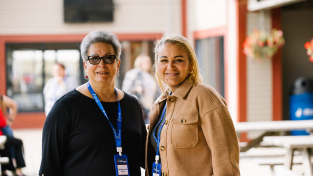 Two people smiling outdoors, wearing name tags and casual attire, with a blurred building and people in the background.