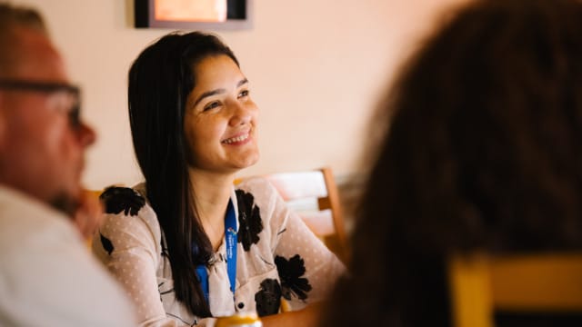 Smiling woman in a meeting or group discussion setting, seated at a table, with a beverage can in front of her.