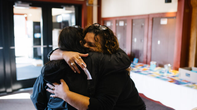 Two people embracing warmly inside a building with tables in the background.