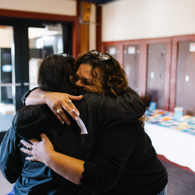 Two people embracing warmly inside a building with tables in the background.