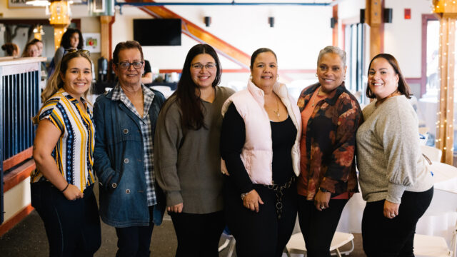 Group of six people standing together inside a warmly lit indoor setting with wooden beams in the background.