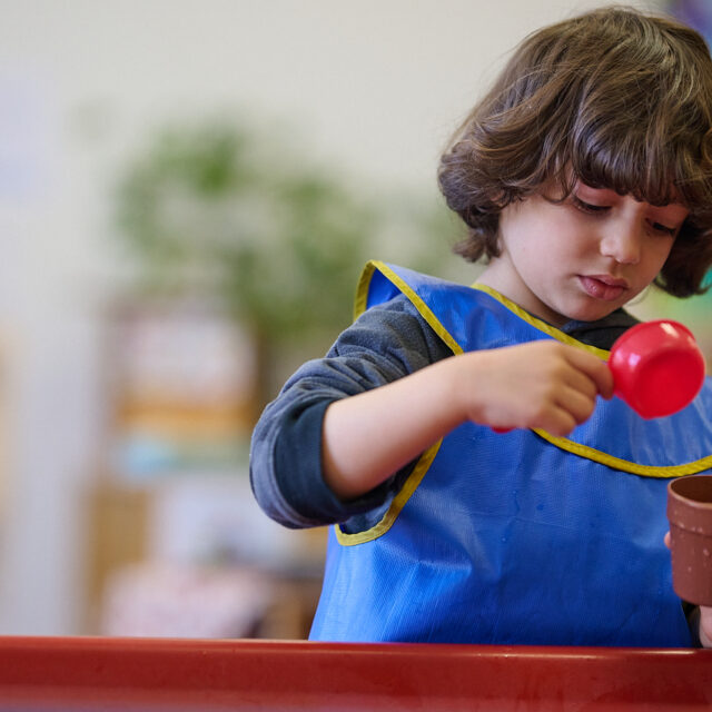 Child pouring water into a small pot wearing a blue apron during a playful activity in a classroom setting