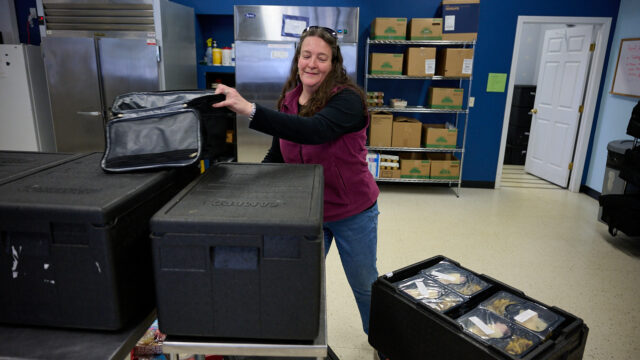 Person organizing packed food containers in a kitchen setting with shelves of boxes and supplies in the background.