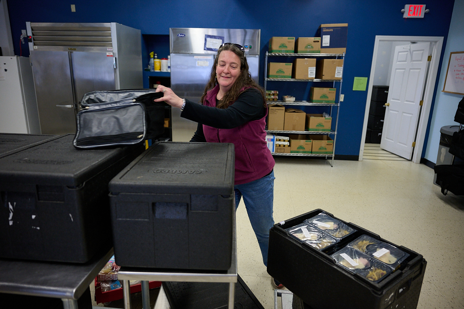 Person organizing packed food containers in a kitchen setting with shelves of boxes and supplies in the background.