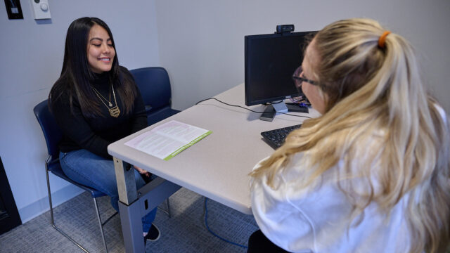Two women sitting at a table in an office setting, one is smiling while the other works at a computer.