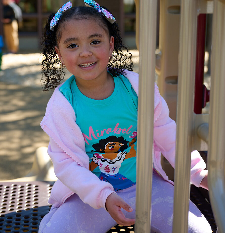 Young girl smiling on playground equipment, wearing a colorful shirt and pink jacket, with curly hair tied in bows.