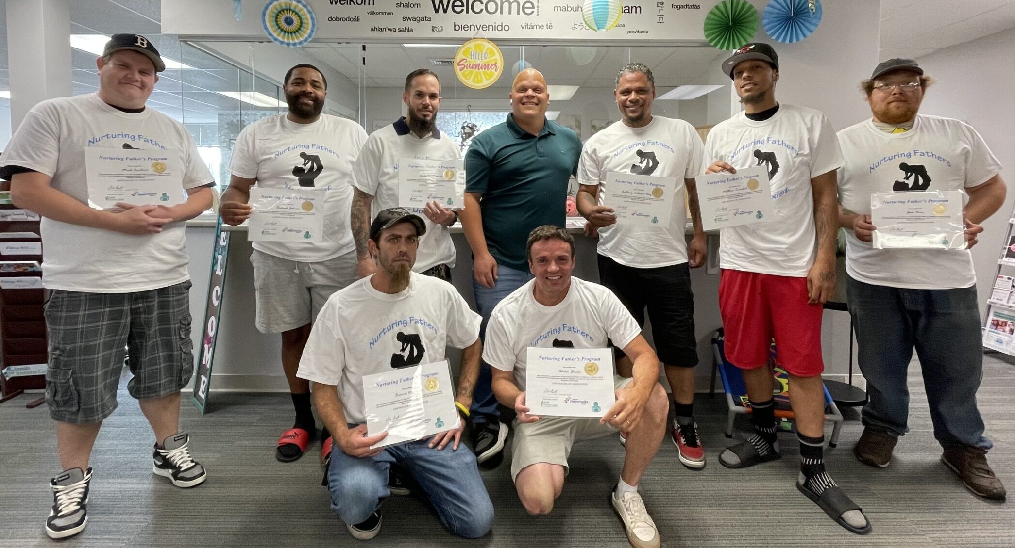 Group of men smiling and holding certificates in a room with welcome banners and colorful decorations.