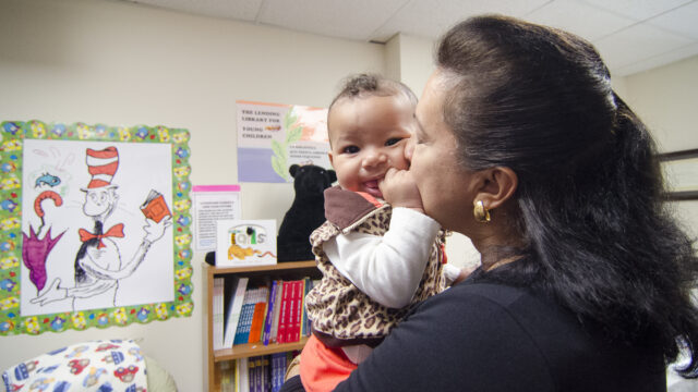Woman holding a smiling baby in a room with colorful posters and bookshelves.