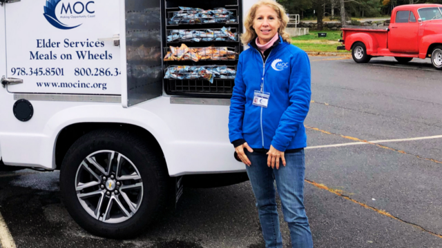 Person in blue jacket standing next to a white truck with open door displaying meal trays for elder services Meals on Wheels.