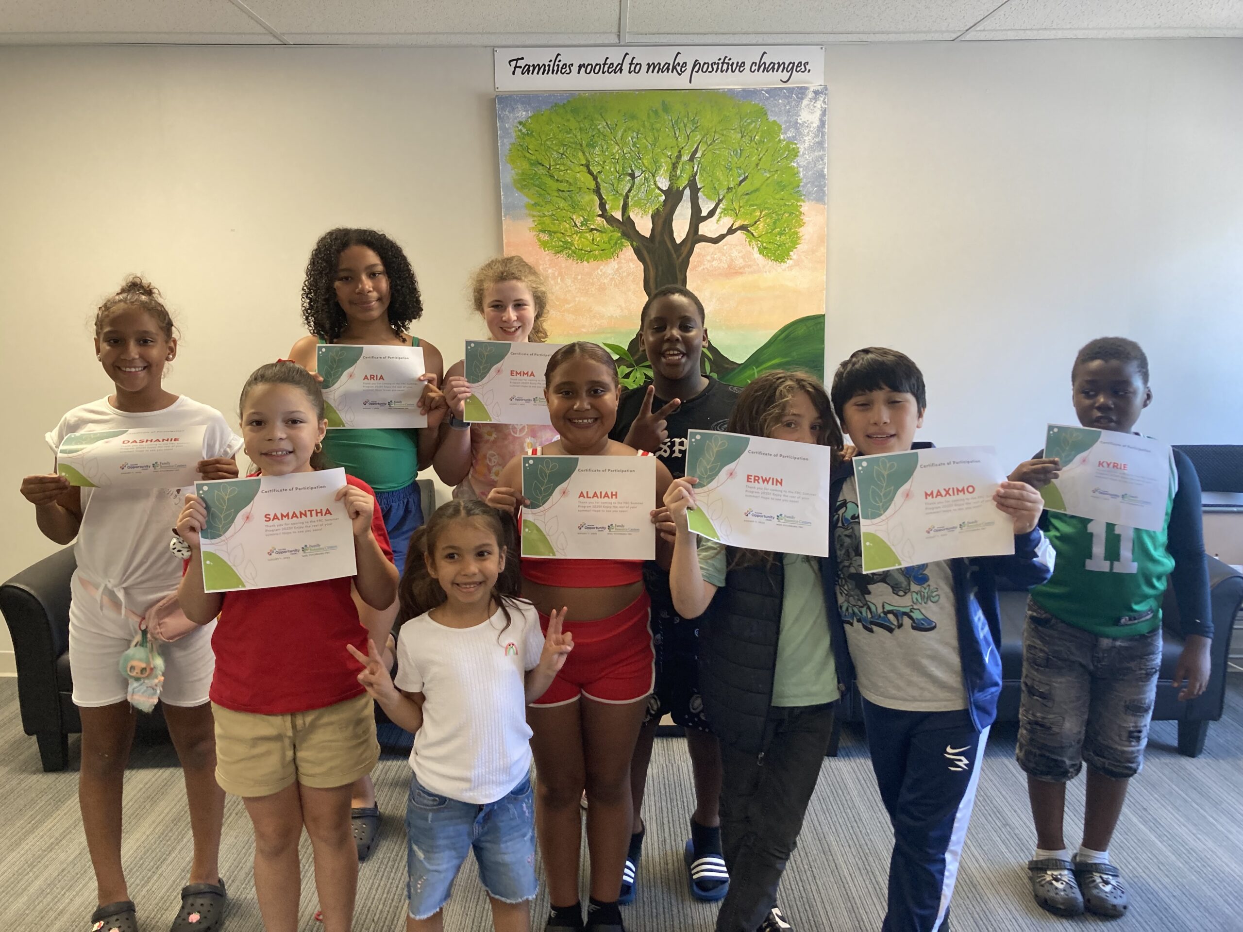 A group of children smiling and holding certificates in front of a mural with a tree.
