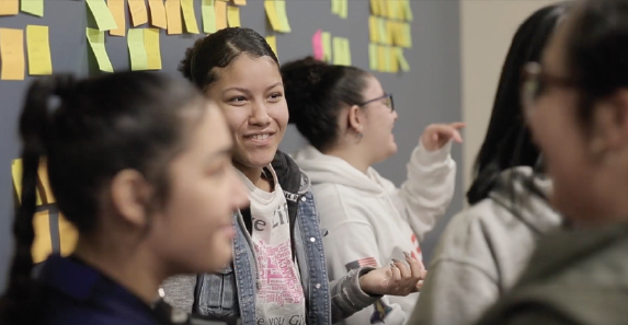 Students engaging in a lively discussion, with colorful sticky notes on the wall in the background.
