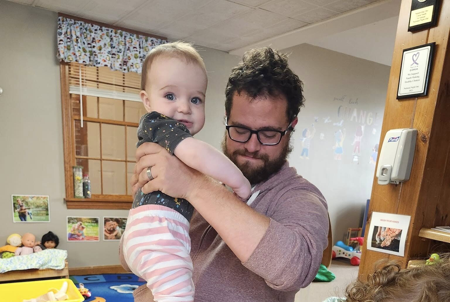 Man holding baby while another child colors paper cutouts at a table in a playroom.