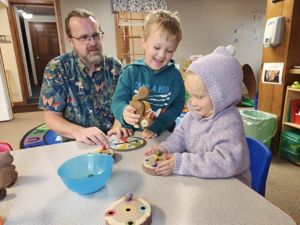 Man with two children playing with animal toys at a table in a colorful room.