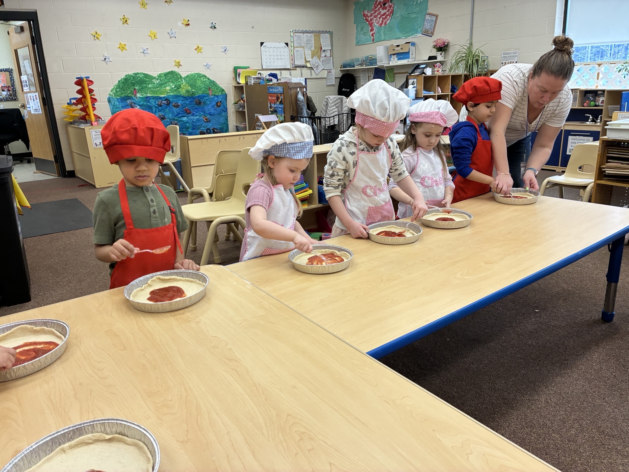 Children in chef hats and aprons making pizzas at a classroom table with the help of an adult.