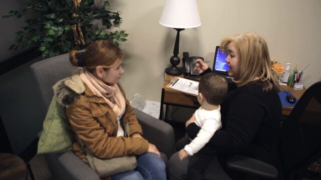 Counselor with child and parent in office, discussing and pointing at a tablet on the desk with a form and lamp nearby.