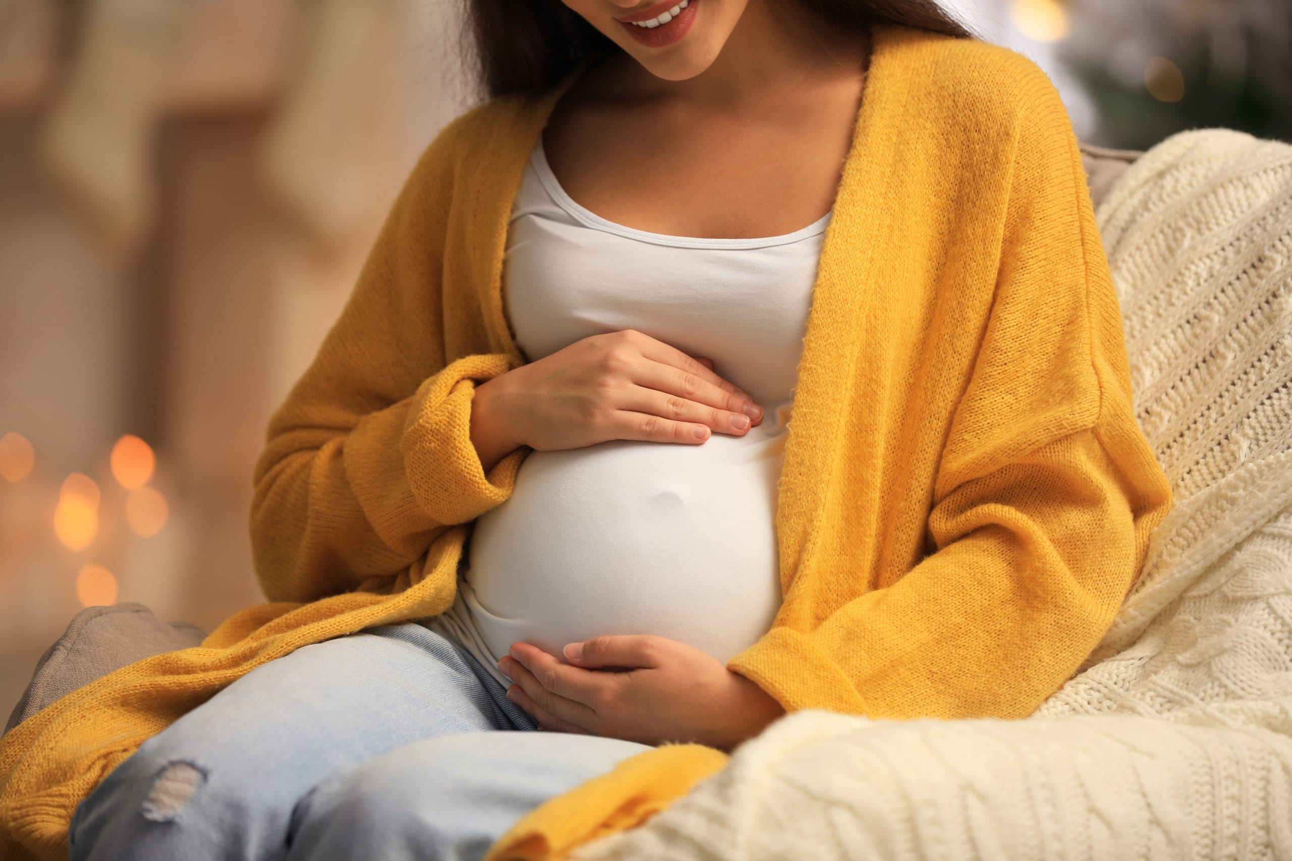 Pregnant woman smiling and cradling her belly, sitting on a cozy couch with warm lighting and a yellow cardigan.