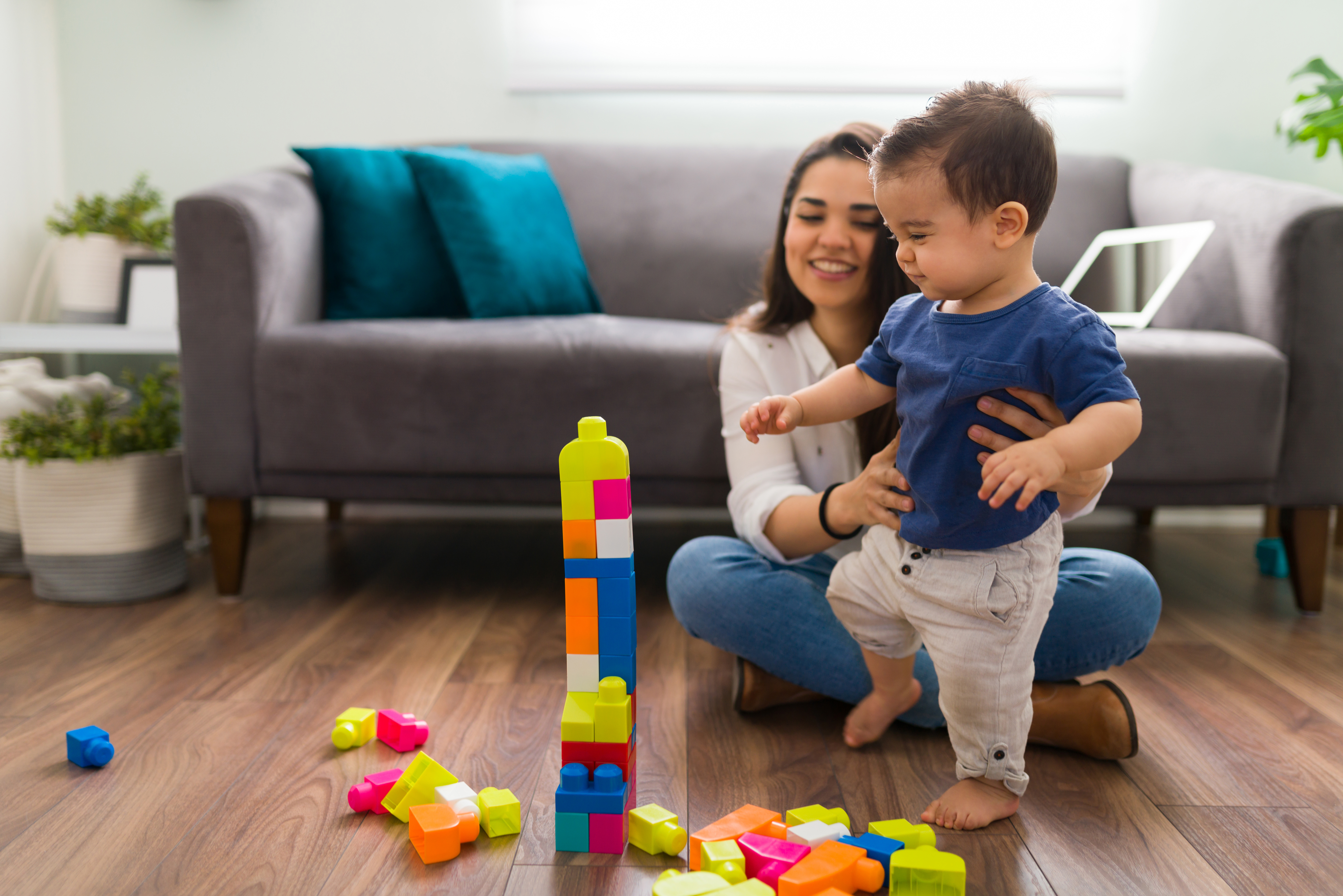 A smiling woman helps a toddler stand by colorful building blocks in a cozy living room.