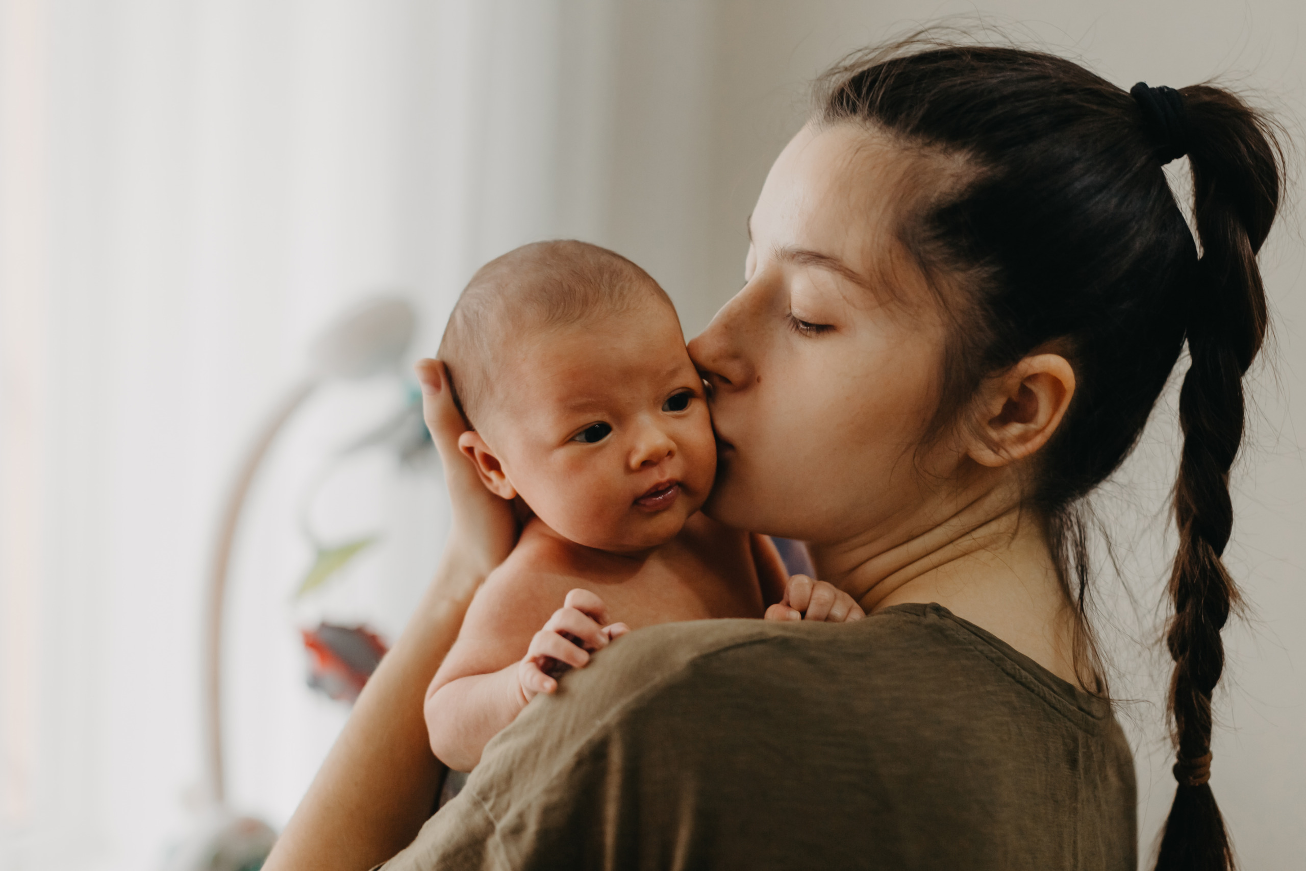 Mother lovingly kisses her newborn baby, who is resting calmly in her arms, near a softly lit window.