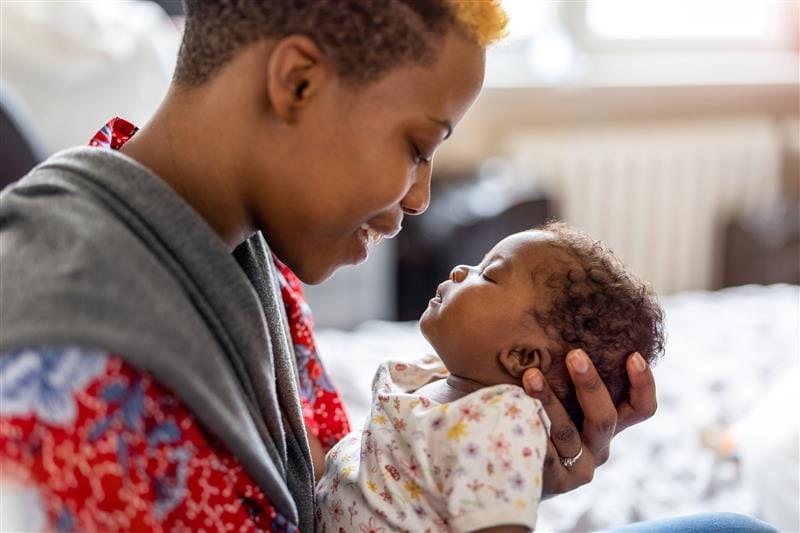 Parent holding a sleeping baby, both smiling softly in a cozy room.
